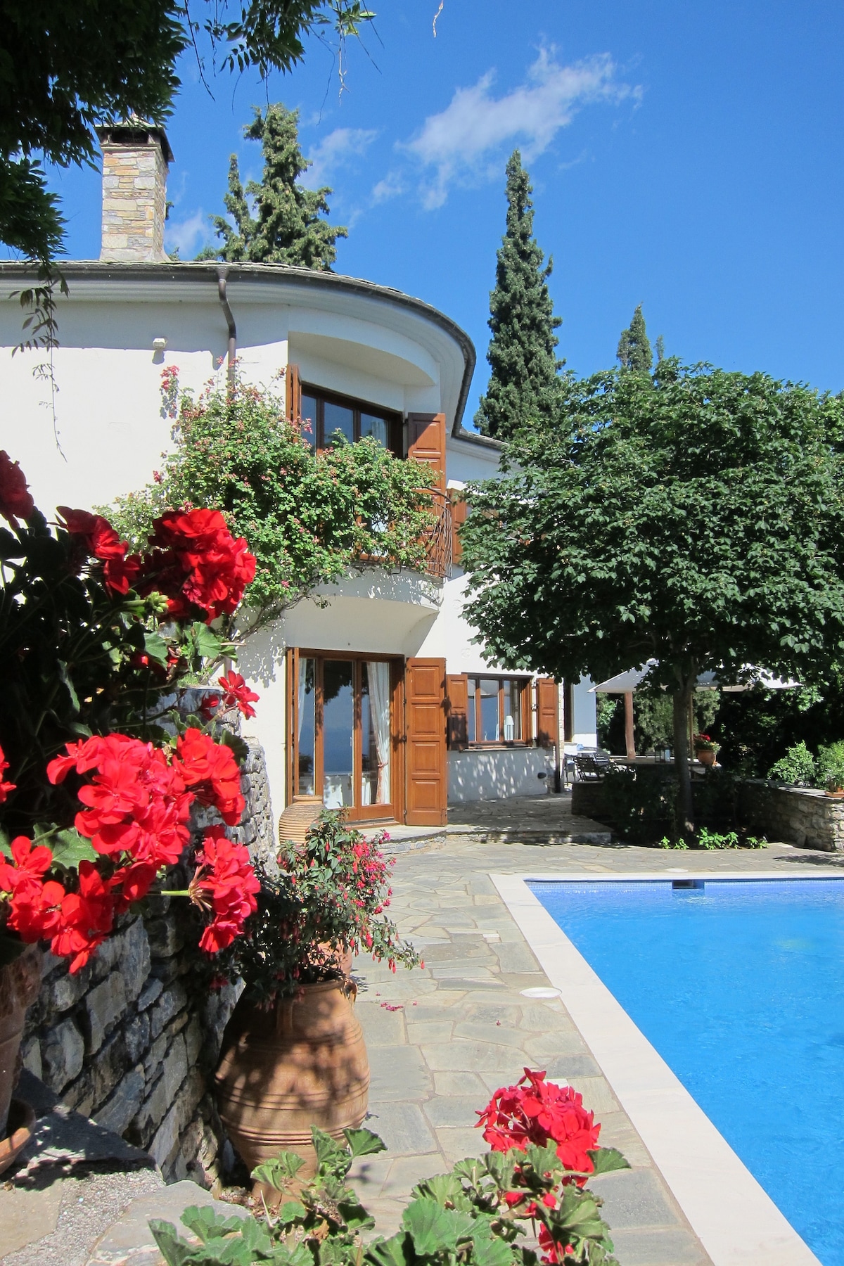 The villa's exterior features vibrant red flowers in terracotta pots, complemented by a clear blue swimming pool. Large windows framed by wooden shutters reflect the sunny sky, while tall trees provide a natural backdrop. The stone pathway leads towards a welcoming entryway.