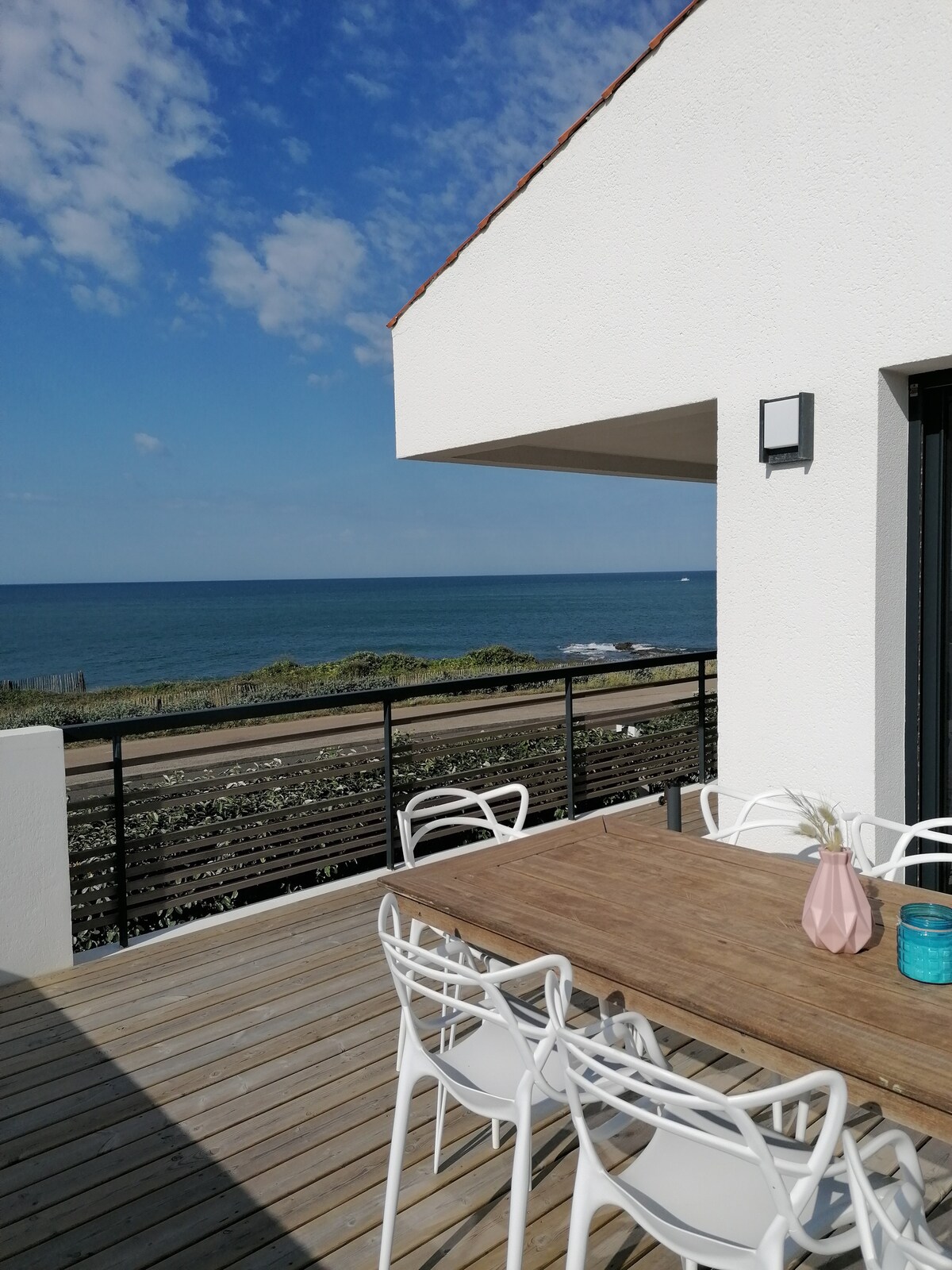 An outdoor terrace features a spacious wooden dining table accompanied by six white chairs. The sea is visible in the background, extending under a clear blue sky. Greenery lines the coastline, emphasizing the tranquil setting.