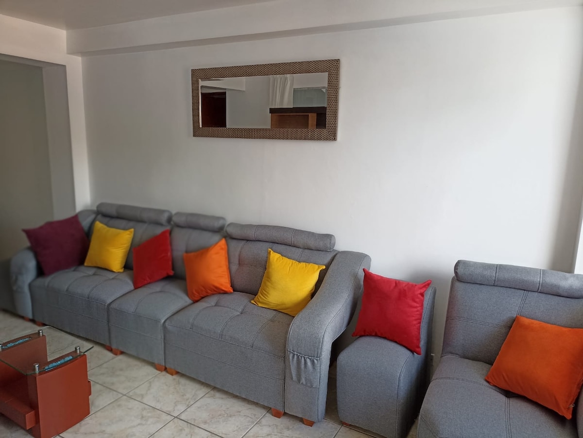 A comfortable living area is shown with a gray sectional sofa featuring vibrant cushions in red and yellow. A mirror reflects the light, enhancing the open feel of the space. Tiled flooring complements the furniture arrangement.