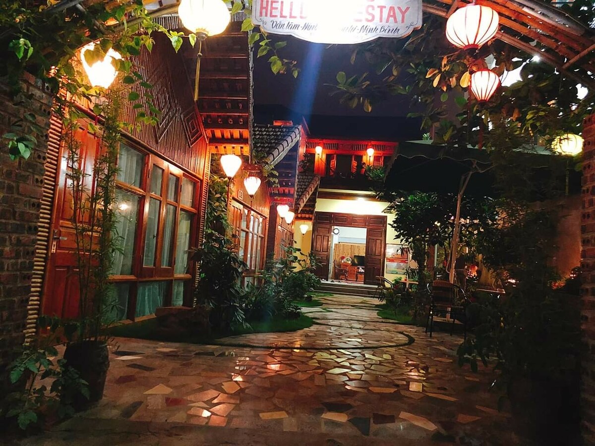 A warm courtyard is illuminated by decorative lanterns, casting a soft glow on the stone pathway. Lush greenery and potted plants are integrated throughout the space, framing the entrance to the bungalow. Windows line the walls, adding an inviting feel to the tranquil setting.