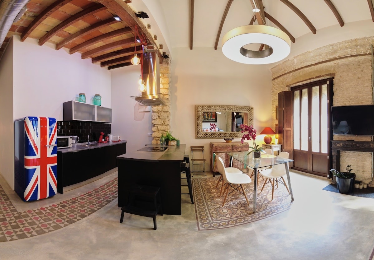 The kitchen features a blend of modern appliances and traditional architecture. A unique British-themed refrigerator adds character, while a spacious island provides additional preparation space. Stylish dining chairs are arranged around a small table, and large windows allow natural light to fill the area.