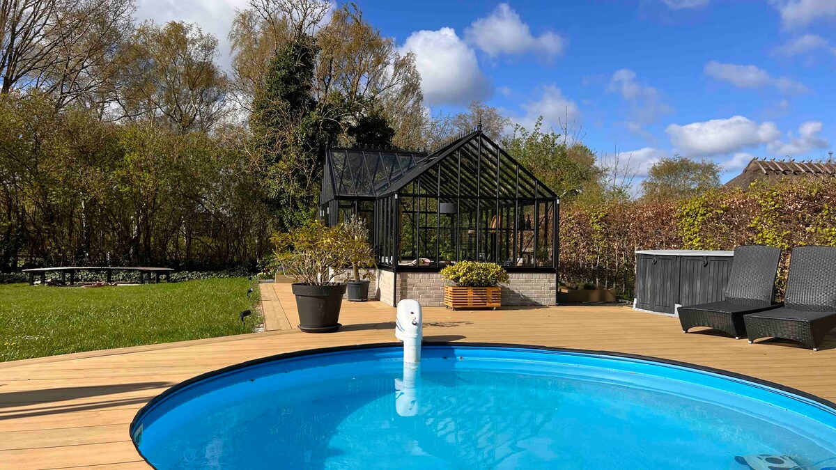 A clear blue pool is surrounded by wooden decking, with lounge chairs positioned nearby. A glass greenhouse is visible in the background, framed by lush greenery. The sky is bright with a few scattered clouds, enhancing the outdoor space's inviting feel.