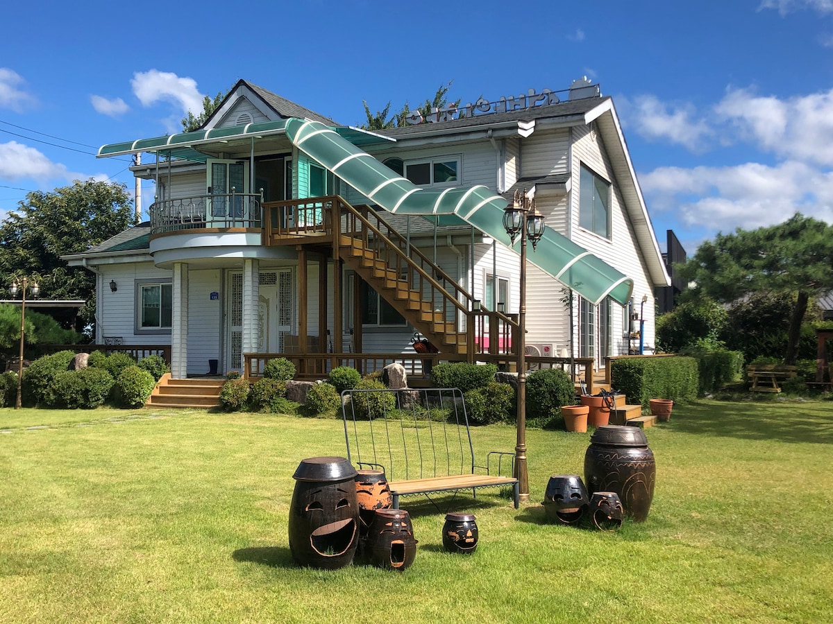 A two-story house features a spacious porch with a green awning and wooden staircase leading to the entrance. The surrounding lawn is well-maintained, showcasing decorative pots and a wooden bench, while lush greenery frames the property under a clear blue sky.