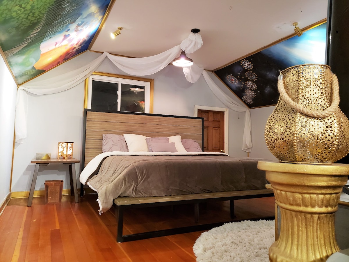 The upper-level bedroom features a spacious king-size bed with a memory foam mattress, complemented by decorative murals on the ceiling. A cozy area rug lies on the hardwood floor, while soft lighting and elegant furnishings enhance the room's unique character.