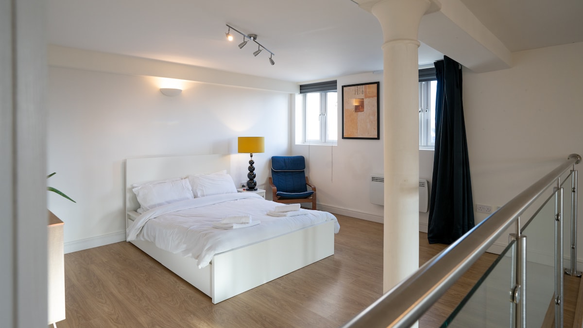 A spacious bedroom is situated on the mezzanine level, featuring a light-colored bed with fresh linens. A comfortable armchair is placed nearby, and light enters through two windows that are fitted with blinds. A lamp on a bedside table adds to the functionality of the space.