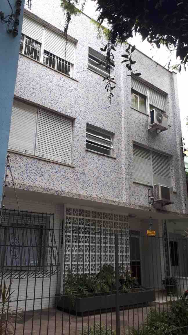 An exterior view of a multi-story building is presented, featuring a façade adorned with a textured pattern. Several windows equipped with protective shutters are visible, along with air conditioning units. A small garden area with greenery is located at the entrance, and a gated fence surrounds the property.