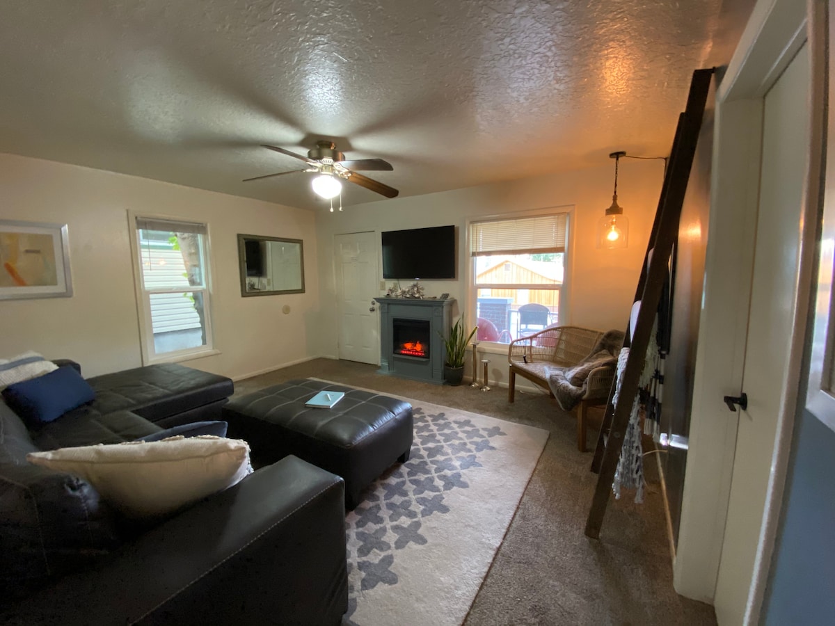 A comfortable family room features a large sectional sofa and a decorative ottoman. A flat-screen TV is mounted on the wall, with a cozy electric fireplace below. Natural light is provided by windows adorned with white curtains, and a ceiling fan offers additional ventilation.
