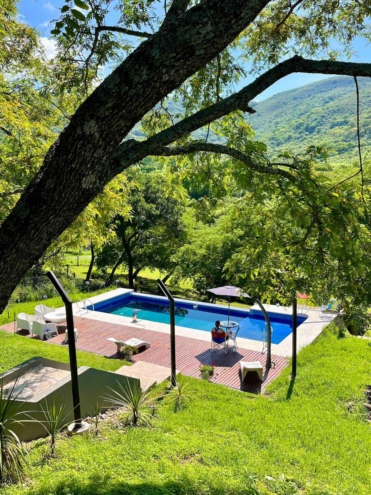 An infinity pool is set amidst lush greenery, offering stunning views of the mountains. Comfortable lounge chairs are positioned around the pool area, creating a serene space for relaxation. A wooden deck enhances the outdoor atmosphere, inviting guests to connect with nature.