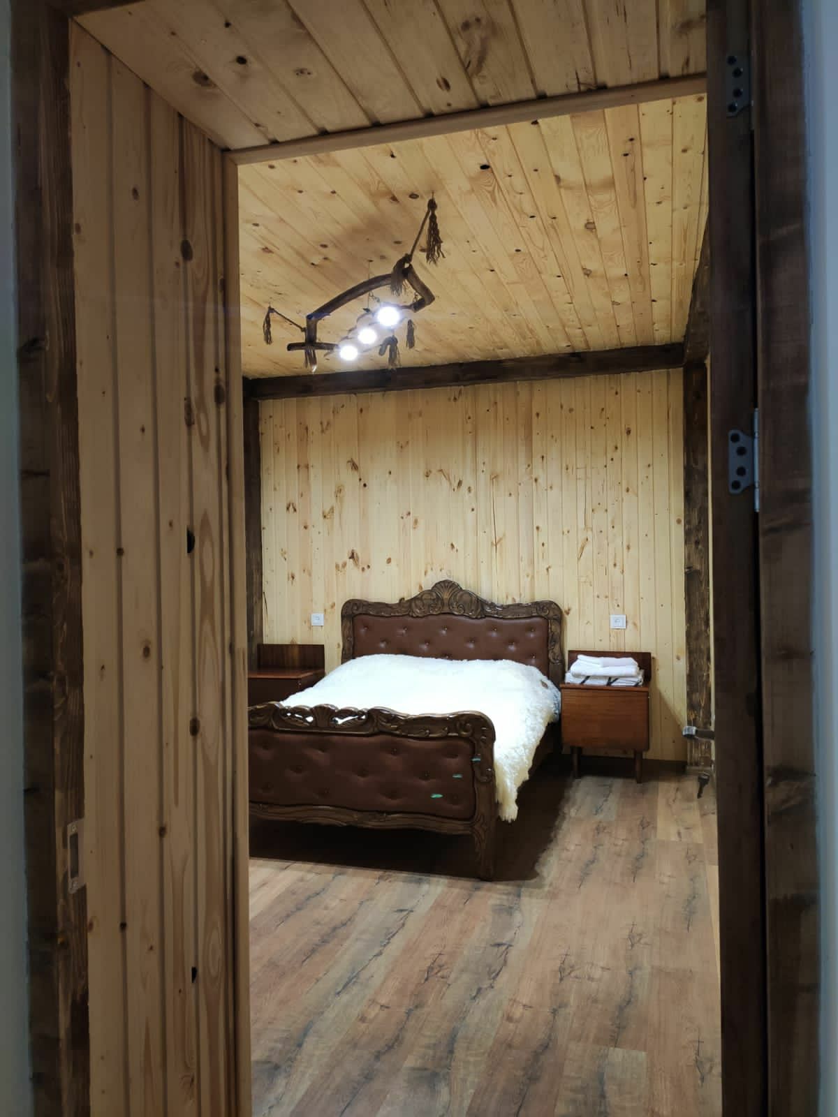 A warm and inviting bedroom is framed by wooden walls and a wooden ceiling. A large, dark wooden bed with a decorative headboard is positioned in the center, complemented by two nightstands on either side. Soft lighting highlights the room's natural materials.