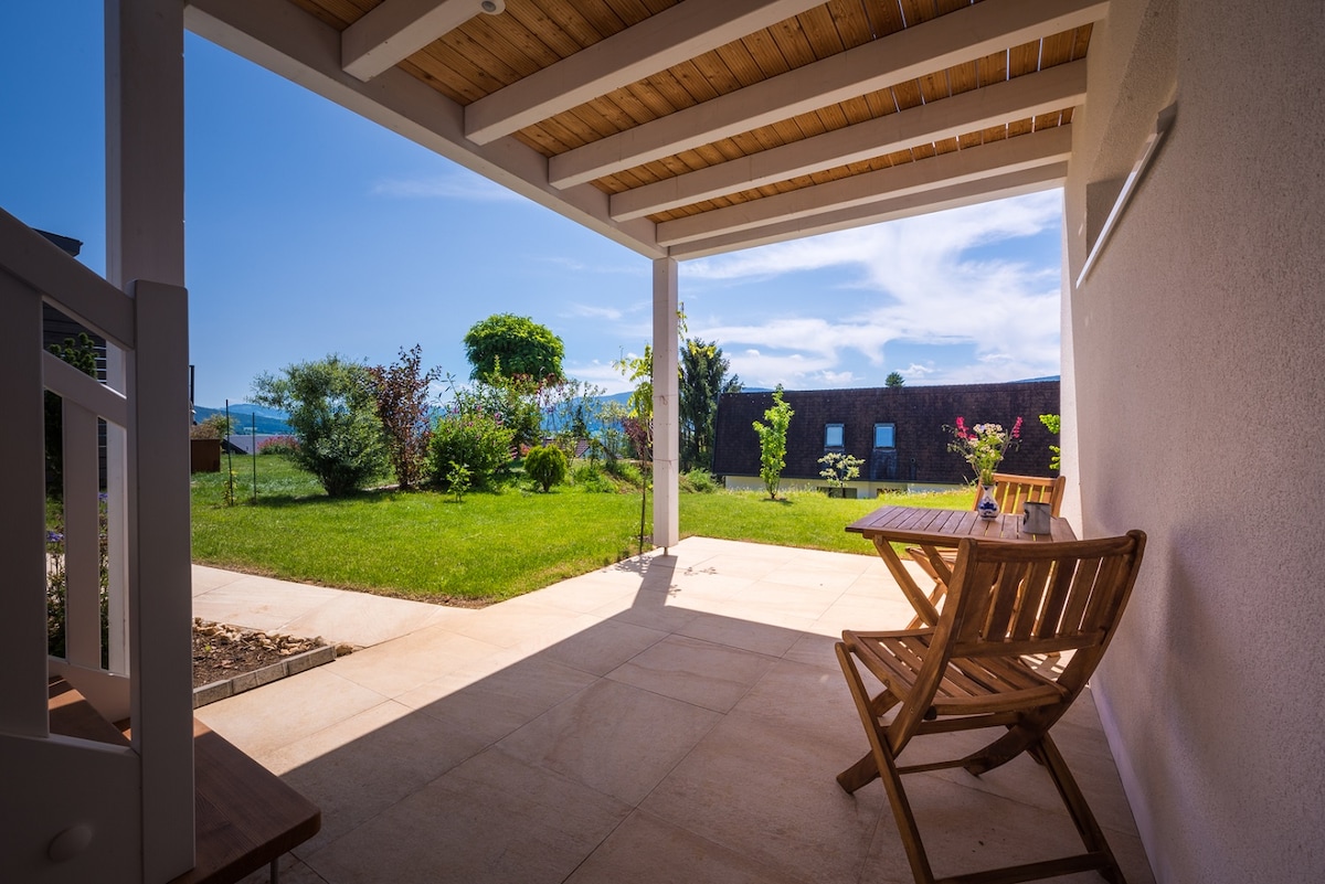 A covered terrace is presented, featuring a wooden table with two chairs. The well-maintained lawn extends into the distance, surrounded by various plants and trees under a clear blue sky, with scenic views of the valley visible beyond.