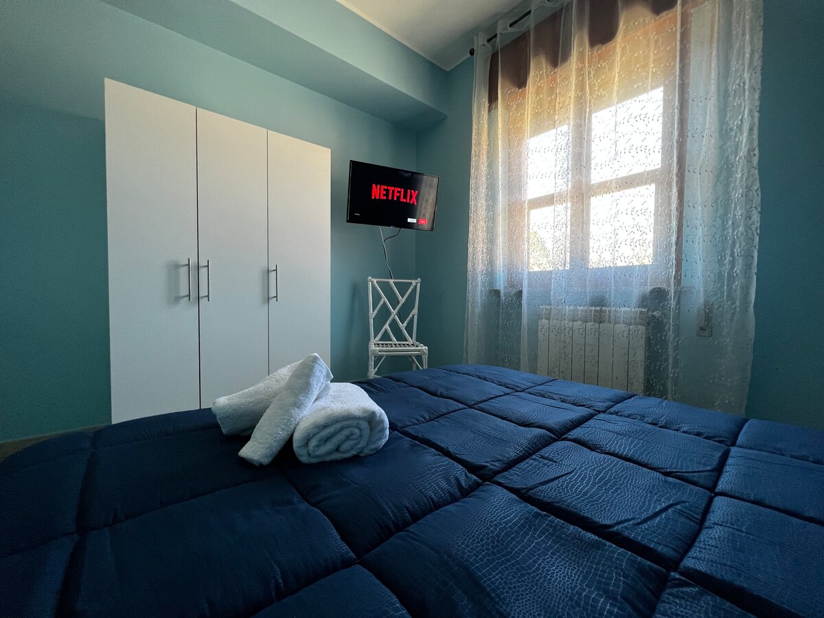 A cozy bedroom features a dark blue quilted bedspread and a medium-sized HD Smart TV mounted on the wall, displaying Netflix. Large windows draped with sheer curtains allow natural light, while a white wardrobe and a chair are positioned against the walls.