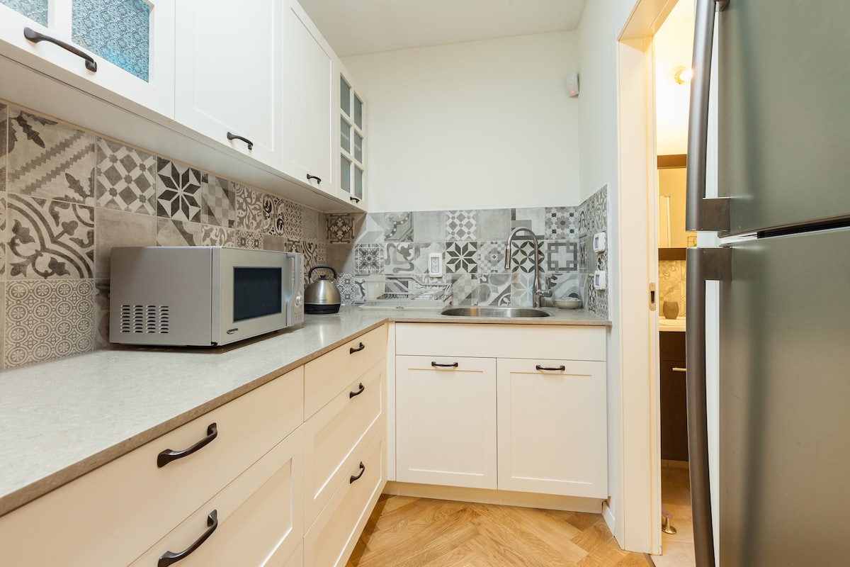 The kitchen area features a modern design with white cabinetry and a patterned backsplash. A countertop and essential appliances, including a microwave and kettle, are visible. Natural light enhances the spacious feel, and the parquet flooring provides a warm touch.