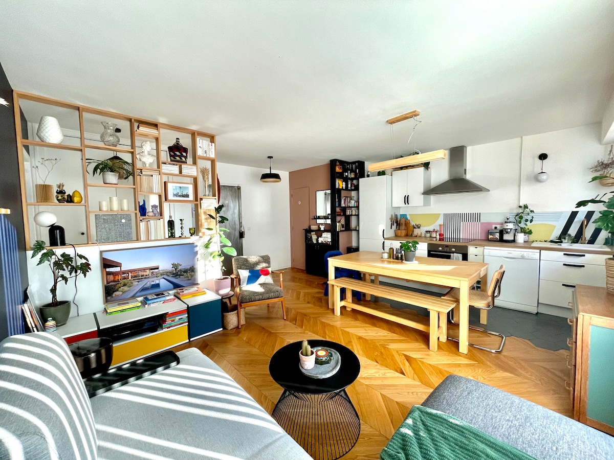 The living area offers a modern and open layout with stylish wooden flooring. A spacious dining table is centrally positioned, surrounded by minimalist decor. A television unit is visible, complemented by shelving displaying decorative items. Natural light fills the room through large windows, enhancing the inviting atmosphere.