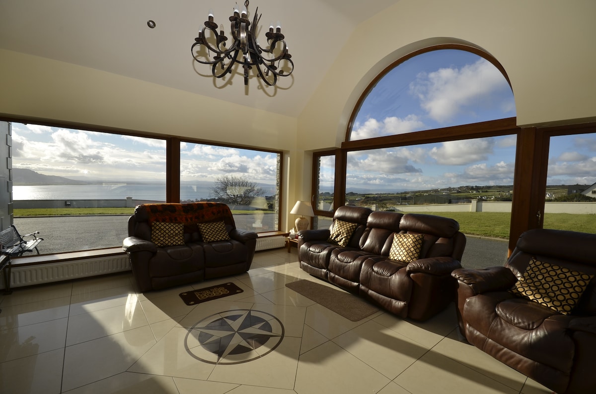 A spacious living area features large arched windows showcasing views of the lough and surrounding landscape. Comfortable leather sofas are arranged around a central decorative rug, and natural light illuminates the glossy tiled floor.