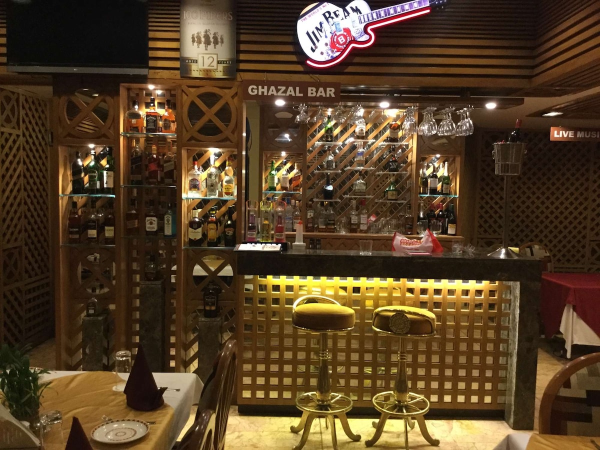 A bar area showcases a wooden design with a guitar-themed sign reading 'Ghazal Bar.' The shelves display a variety of liquor bottles, complemented by glasses hanging overhead. Gold and glass bar stools are positioned in front of a sleek counter illuminated with soft lighting.