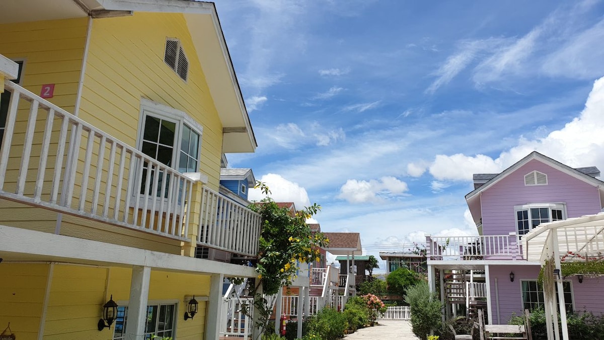 Two colorful, two-story houses are displayed against a bright blue sky, with scattered clouds. The yellow house features a white balcony and lush greenery, while the pink house showcases a charming entrance. Flowering plants line the path leading to both residences.