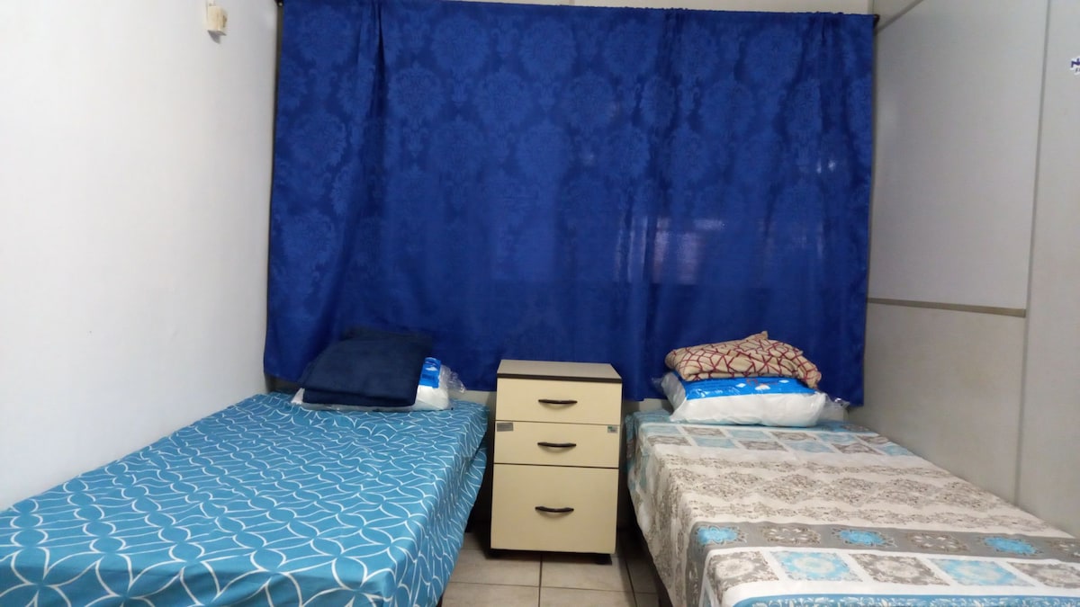 A simple sleeping arrangement is presented, featuring two beds with patterned bedding. A nightstand with three drawers is situated between the beds. A blue curtain covers the window, and neutral-toned tiles are visible on the floor.