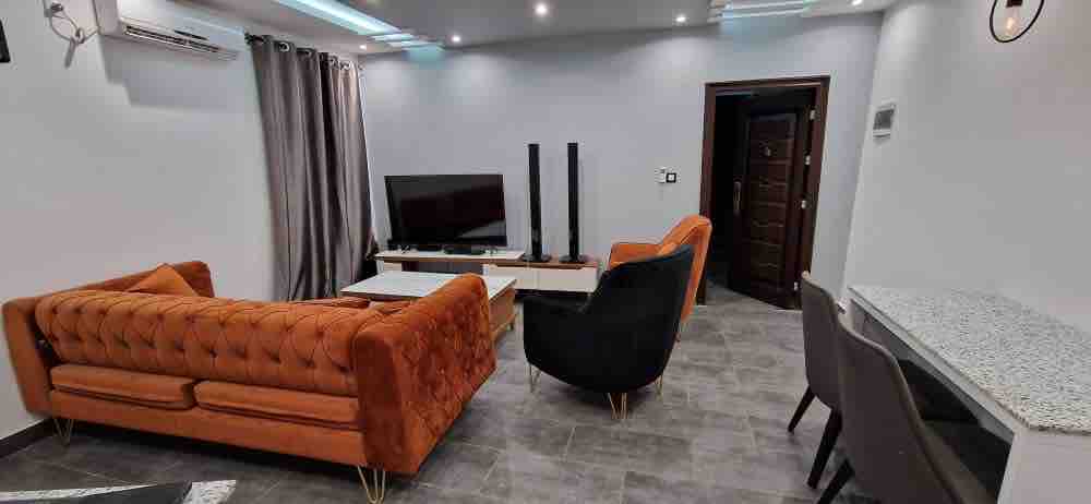 An inviting living area features a stylish orange tufted sofa and a black armchair, set against a neutral wall. A modern coffee table sits in front of a sleek entertainment unit that holds a television. Recessed lighting enhances the overall ambiance.