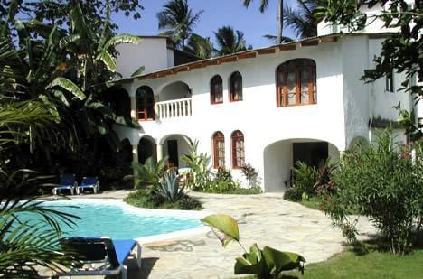 A well-maintained exterior features a white multi-level building surrounded by lush greenery. A clear pool is bordered by patio spaces, with lounge chairs positioned nearby. Arched windows and balconies add architectural interest to the façade, creating a welcoming outdoor environment.