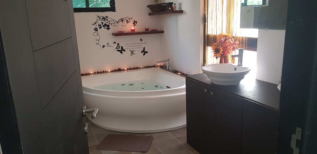 The bathroom features a large, oval jacuzzi tub surrounded by soft lighting and decorative accents. A modern sink is situated on a dark countertop, complemented by a minimalist decor. Natural light filters through elegant curtains, enhancing the serene ambiance.
