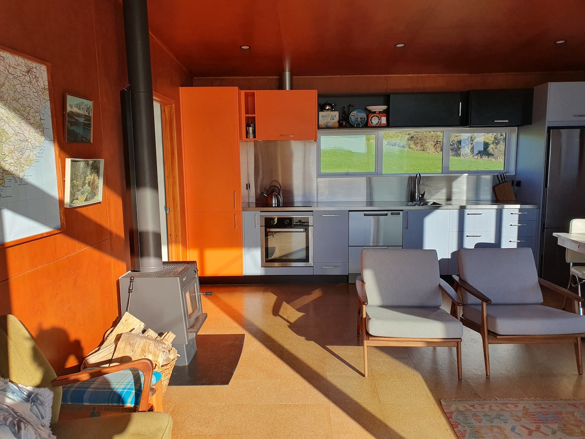 A modern kitchen and living area are displayed, featuring warm orange and cool blue cabinetry. A wood-burning stove adds warmth, and two armchairs offer comfortable seating. Sunlight streams in through large windows, highlighting the spacious layout and views of the surrounding landscape.