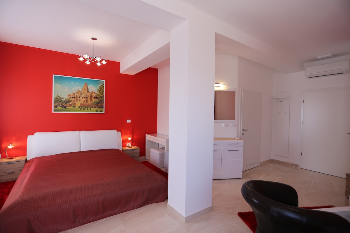 A well-appointed guestroom features a spacious bed covered in rich red bedding, against a backdrop of a vibrant red accent wall. Natural light is provided by a window, and a stylish black chair and modern furnishings complete the elegant atmosphere.