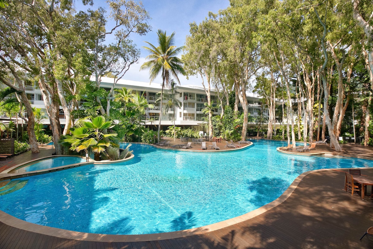 A large lagoon-style swimming pool is surrounded by lush tropical landscaping and tall trees. Sun loungers are positioned around the pool area, providing shaded spots for relaxation. The serene water reflects the sky, creating a tranquil atmosphere for guests to enjoy.
