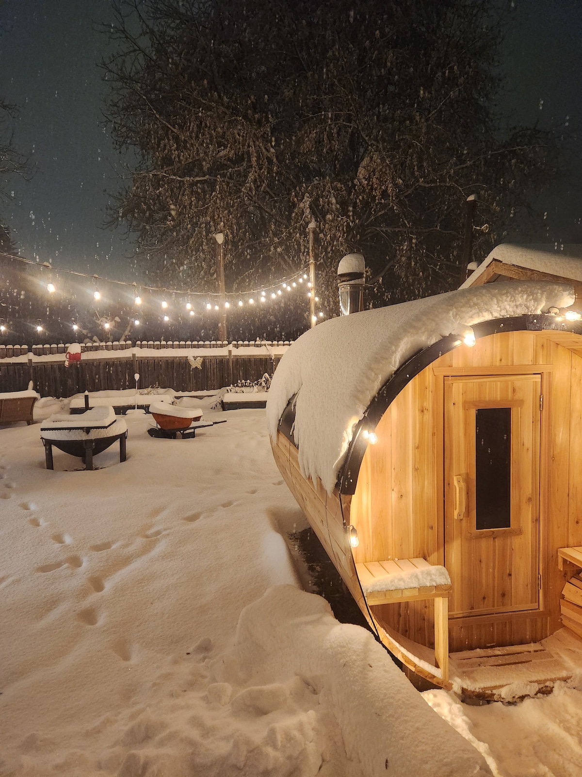 A wood-fired barrel sauna is featured in a snowy outdoor setting, surrounded by soft glowing string lights. A fire pit is visible nearby, set within a spacious yard blanketed in snow. The ambience suggests a serene retreat in a winter landscape.