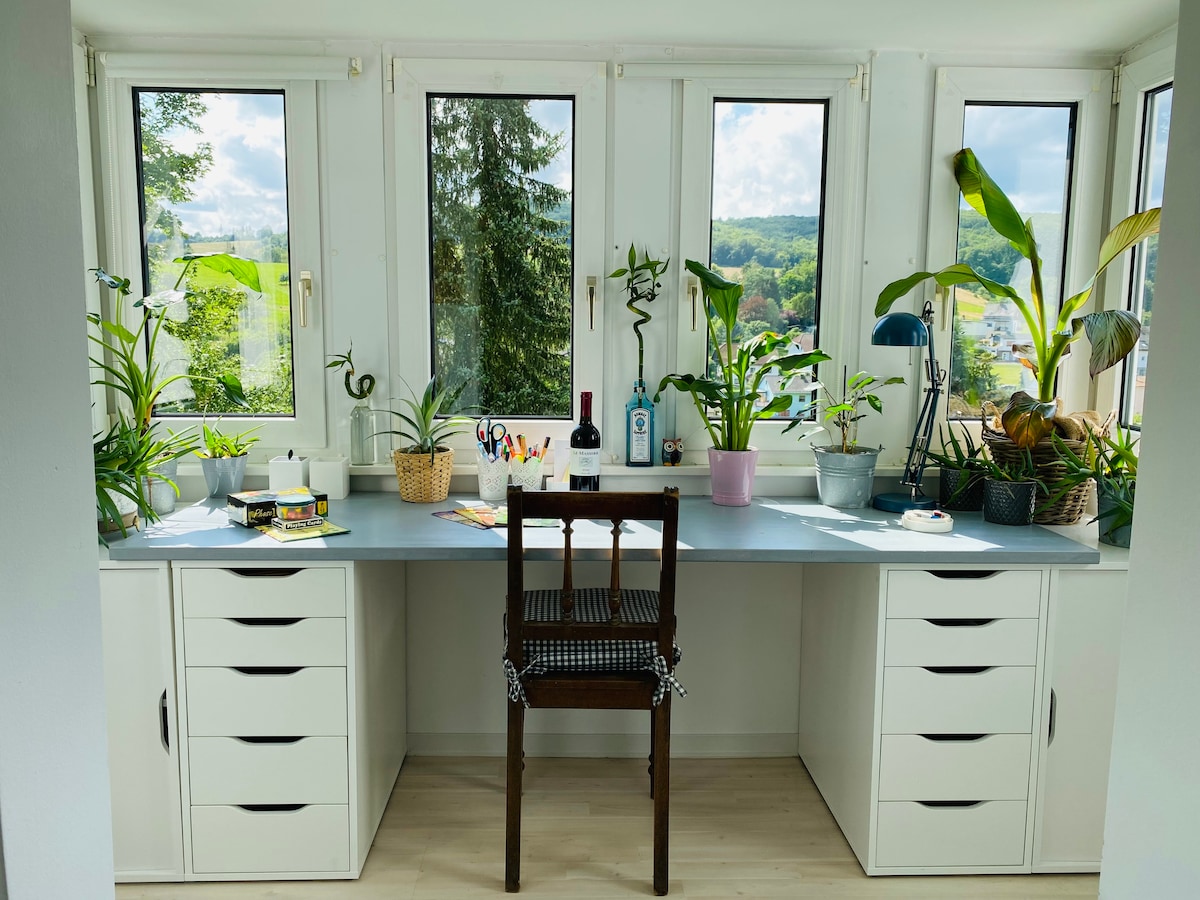 A bright workspace is positioned in front of four large windows, offering views of greenery. A sleek desk with multiple drawers is complemented by a wooden chair. Various potted plants are arranged along the windowsill, enhancing the inviting atmosphere.