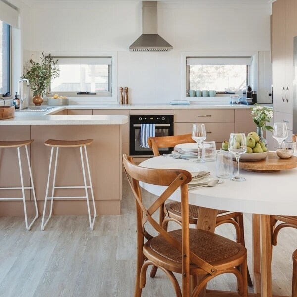 A modern kitchen is presented, featuring light-colored cabinetry and a spacious island with bar seating. A dining table is set with tableware and fresh fruit, while natural light streams in through large windows, highlighting the open layout and inviting atmosphere.