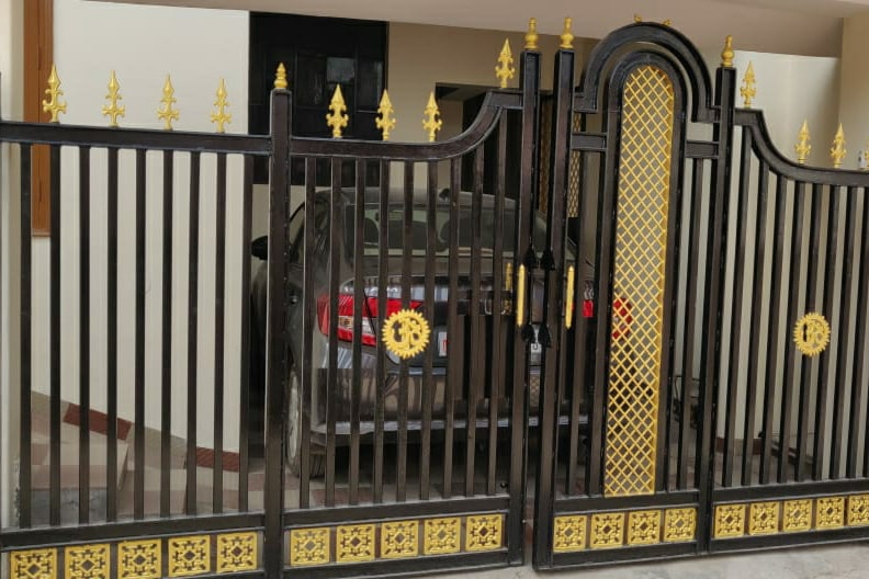 A decorative wrought iron gate features intricate golden designs, providing access to a private parking area. A vehicle is positioned within the enclosed space, indicating availability for guest parking. Neutral wall colors are visible in the background, enhancing the entrance's clean appearance.