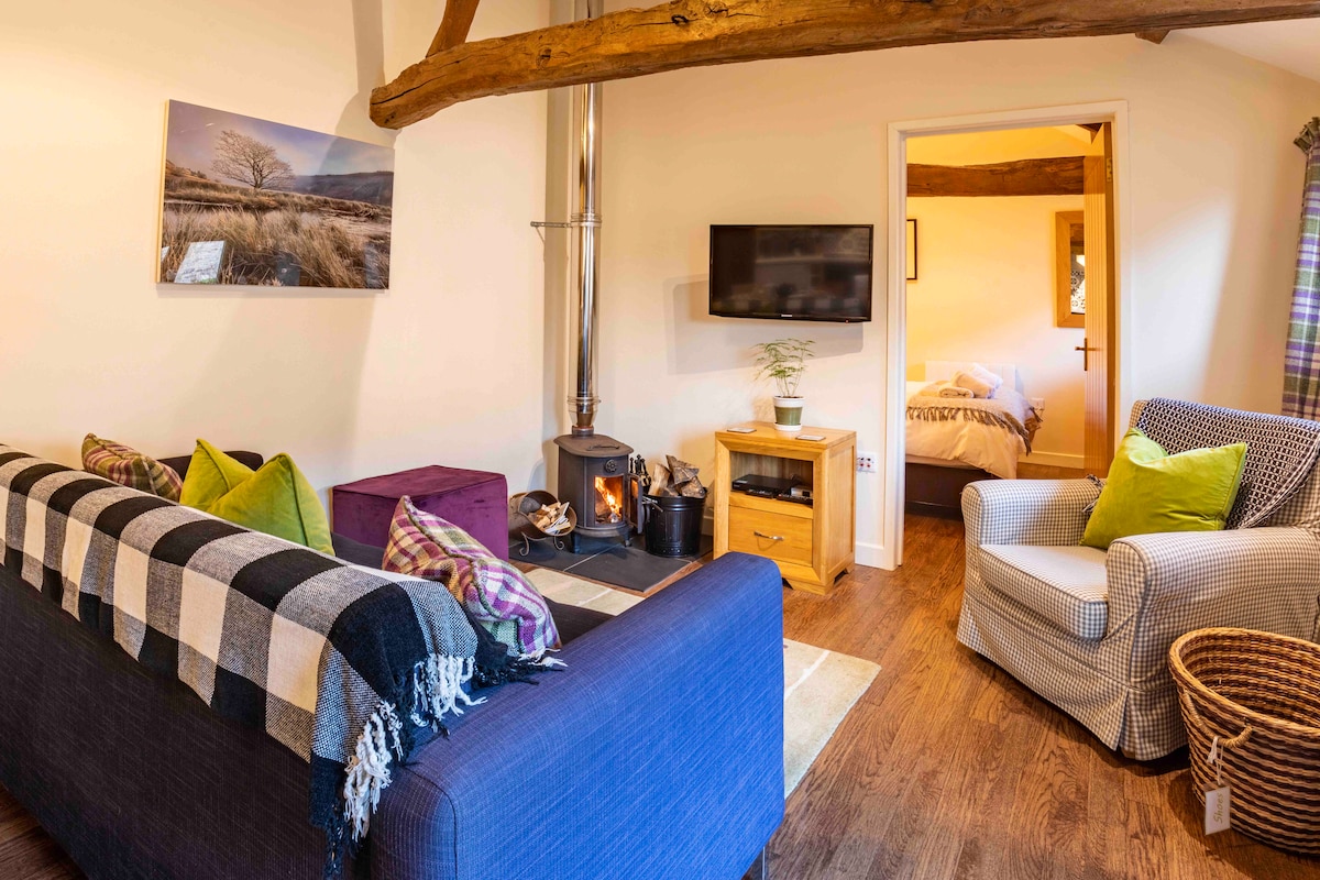 The sitting area features a comfortable sofa adorned with cushions, complemented by an armchair and a wood burning stove nearby. A flat screen television is mounted on the wall, and a door leads to one of the bedrooms, visible in the background.