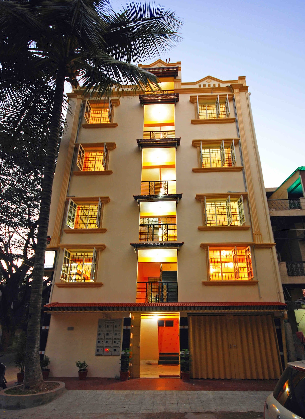 A multi-story building is presented, featuring an array of bright windows that illuminate the exterior. Palm trees are visible nearby, and the entrance is accessible with a covered area for parking. The overall structure is characterized by a clean and inviting design.