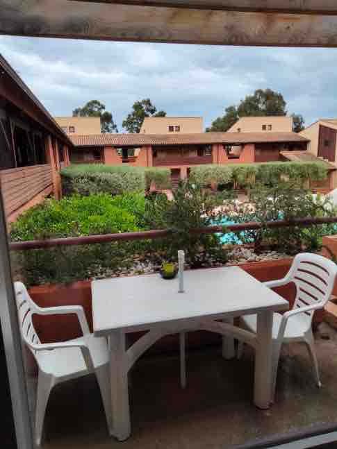 A simple outdoor balcony features a white table and two plastic chairs. Lush greenery and a swimming pool are visible in the distance, framed by the surroundings of the two-story residence. The setting provides a serene view for relaxing moments.
