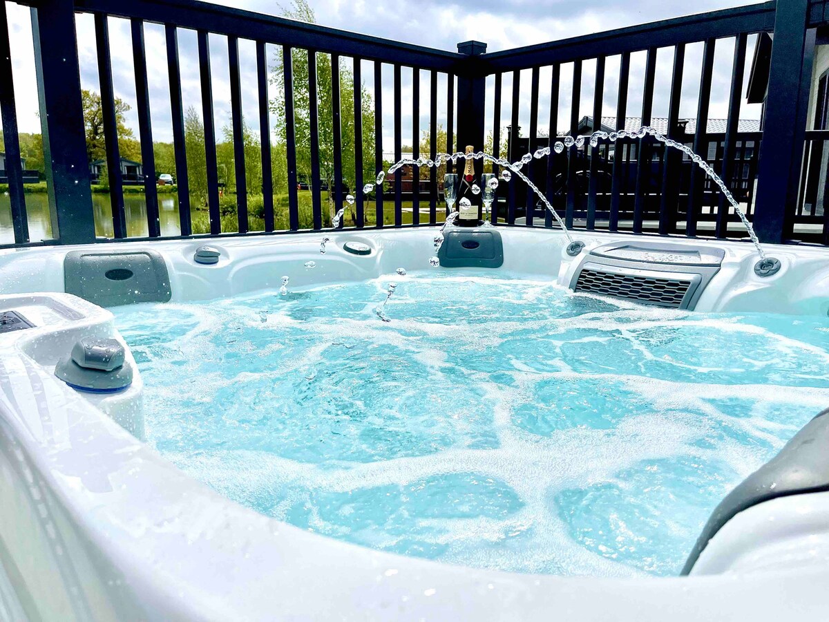 A luxury hot tub features bubbling water and several water jets, surrounded by a black deck railing. The serene lake setting can be glimpsed in the background, contributing to a relaxing outdoor atmosphere.
