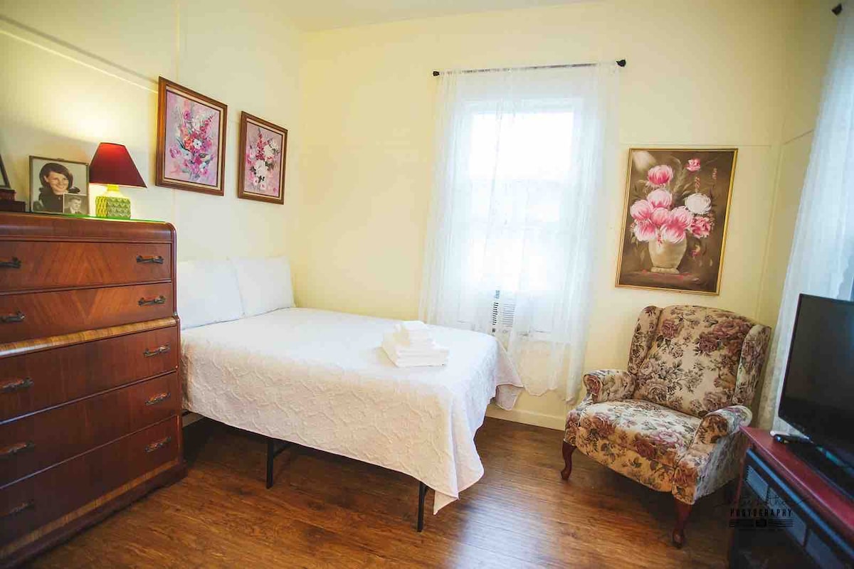 A bright and airy sleeping space features a cozy bed with a white quilt and neatly arranged towels. Vintage floral artwork is displayed on the walls, complementing the wood dresser and a comfortable upholstered chair. Natural light filters through the sheer window curtains.