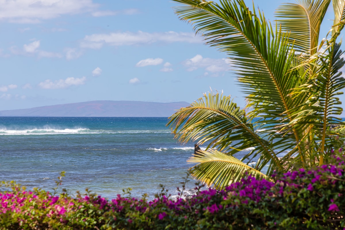 A view of the ocean showcases gentle waves lapping at the shore. In the foreground, vibrant purple flowers and lush palm leaves provide a natural frame, while the distant landmass adds depth to the scenic ocean backdrop.