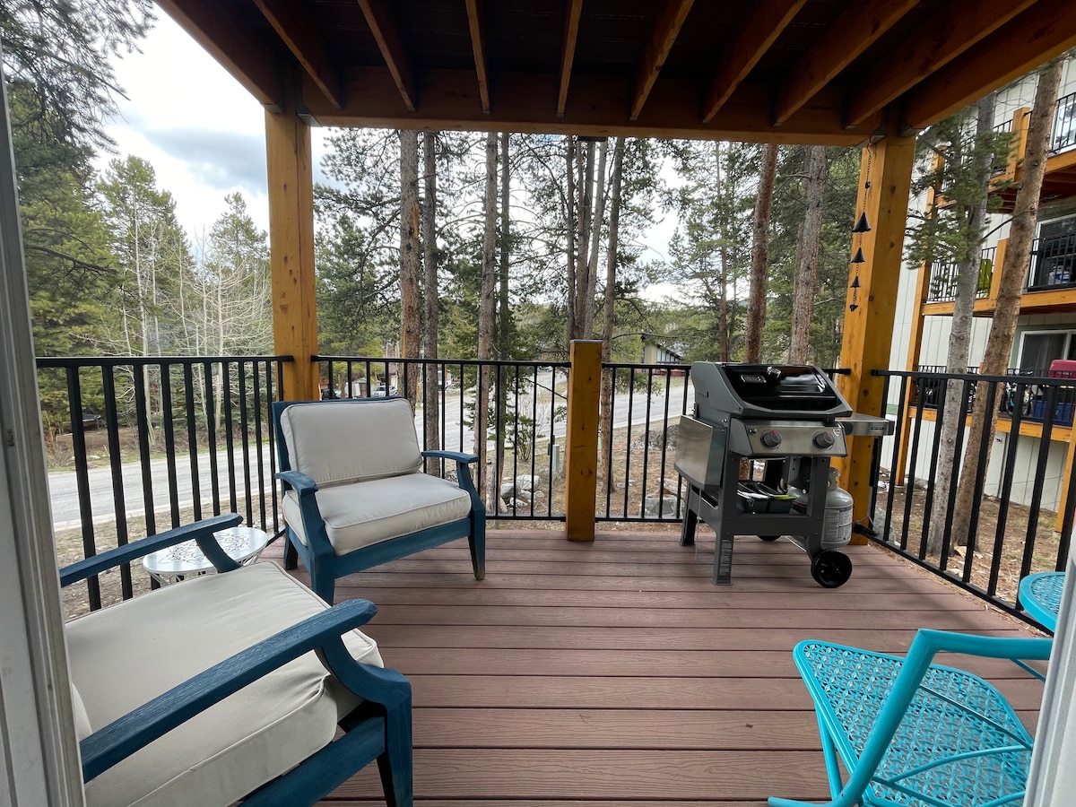 A covered balcony offers outdoor seating with two white chairs and a blue chair. A grill is positioned nearby, surrounded by trees, providing a natural backdrop. The deck features wooden flooring and a railing that enhances the space’s openness.