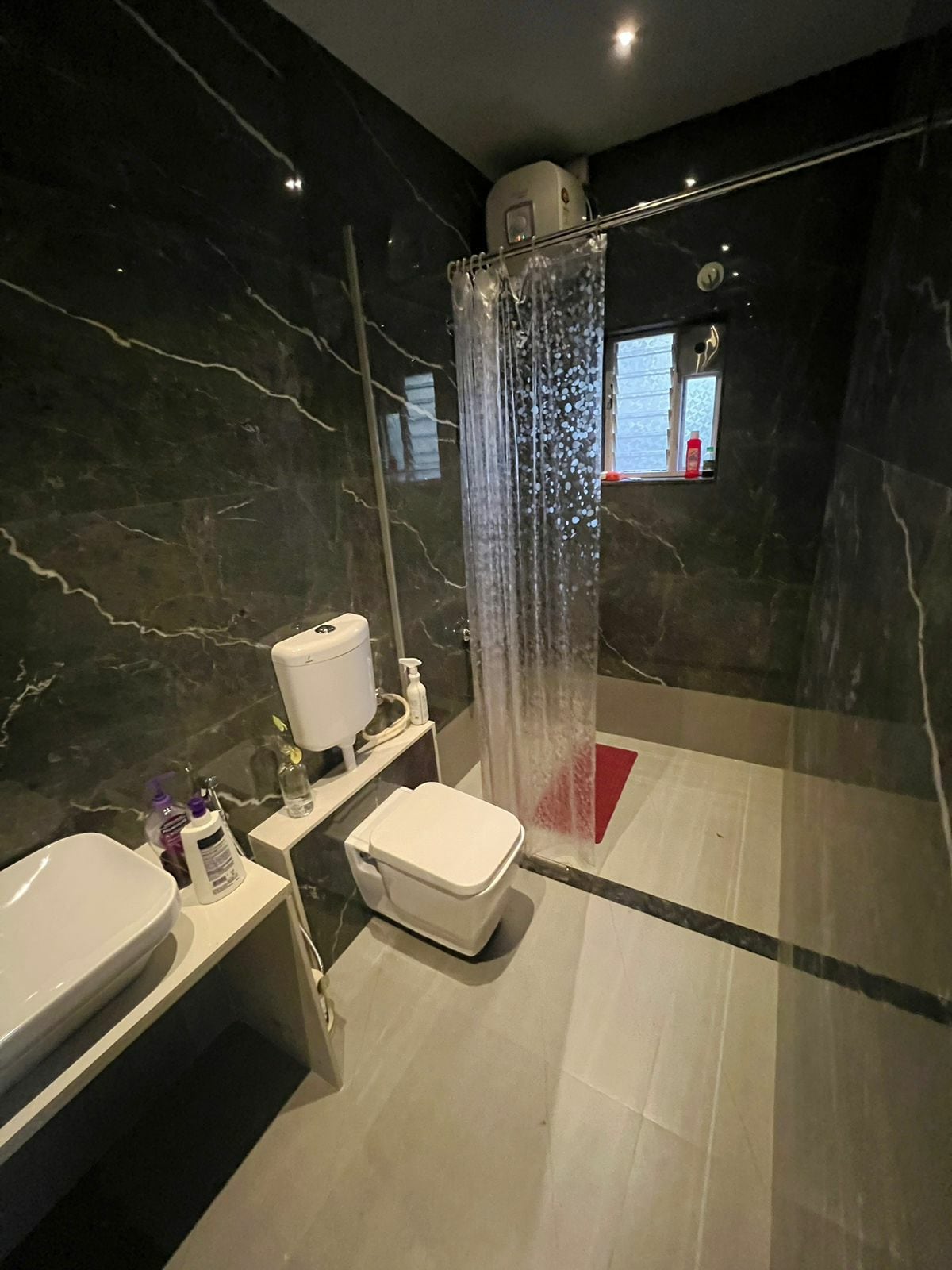 A modern bathroom features marble wall panels and a spacious walk-in shower with a glass partition. A sleek vanity with a wash basin is positioned beside a white toilet. Soft overhead lighting creates a bright ambiance, while a red bath mat provides a pop of color on the floor.