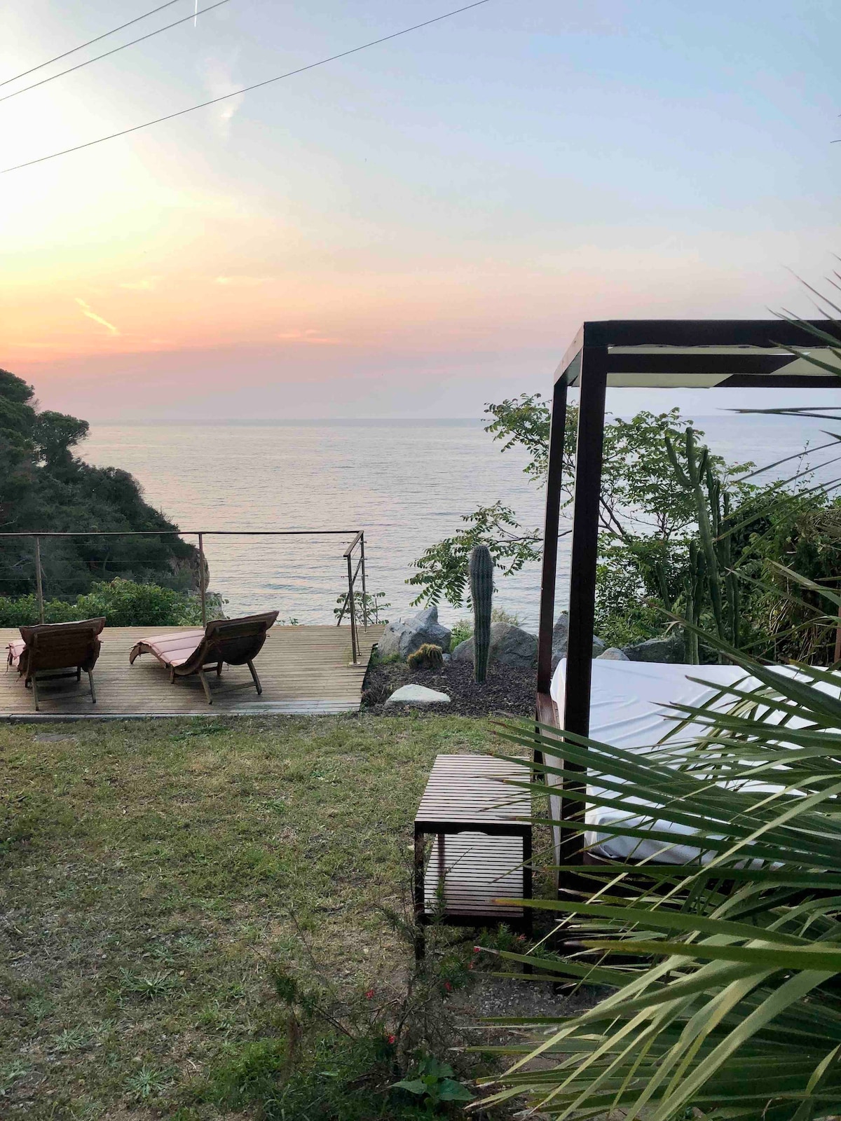 A serene landscape is presented with a view of the sea at sunset, featuring a grassy area with lounge chairs. In the foreground, tropical foliage adds a touch of greenery, while a wooden deck extends towards the water, creating an inviting space for relaxation.