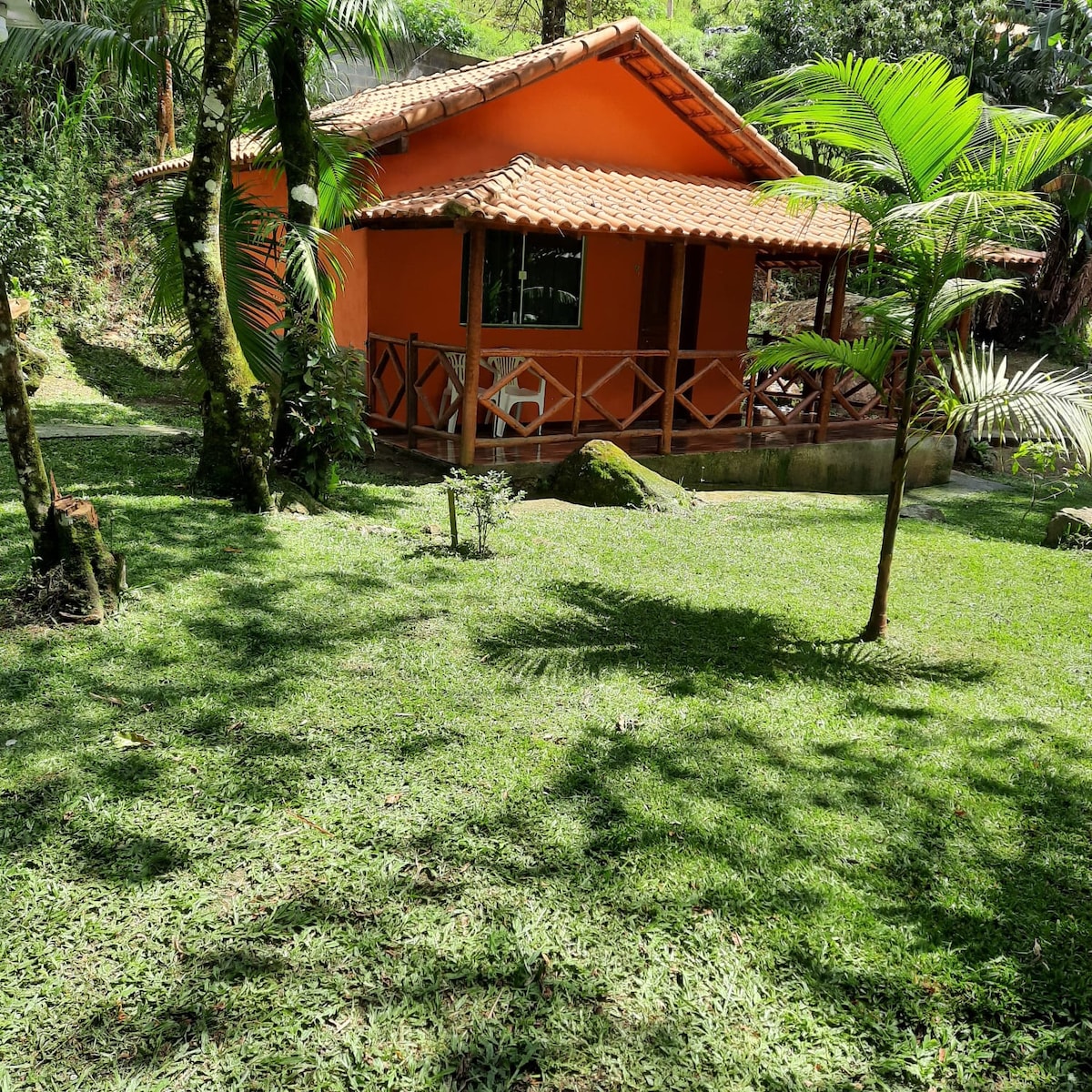 The exterior of the chalet is highlighted, showcasing its vibrant orange façade. Lush greenery surrounds the structure, with a well-maintained lawn and tropical plants enhancing the natural setting. A small patio area with a table is visible, inviting relaxation in the tranquil surroundings.