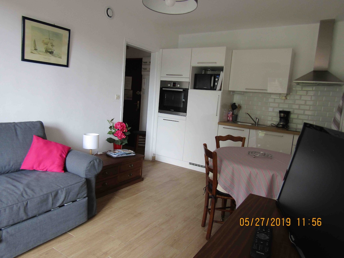 A modern living space is presented, featuring a gray sofa with a pink cushion and a wooden table set for dining. The kitchen area showcases white cabinetry and various appliances, including a microwave and coffee maker. Natural light illuminates the room, creating an inviting ambiance.