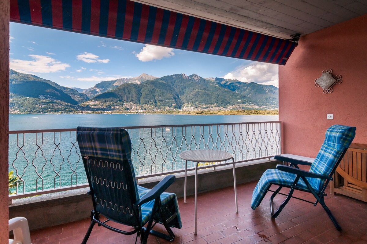 A spacious balcony is featured, showcasing views over Lake Maggiore. Two reclining chairs with patterned fabric are positioned next to a small, round table. A striped awning provides shade, while the tranquil water and distant mountains create a serene backdrop.