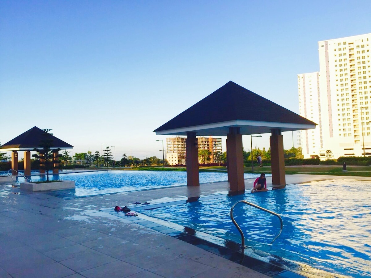 A spacious swimming pool area is surrounded by pavilions with sloped roofs. Lounge chairs are placed nearby, and the water glistens under sunlight. The landscape features green lawns and the silhouette of buildings in the background.