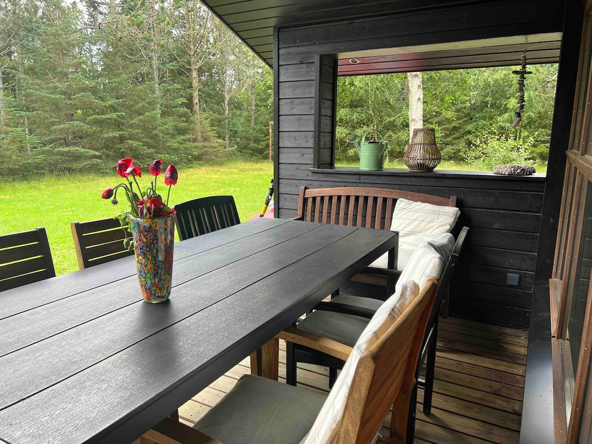 A covered outdoor terrace is depicted, featuring a long black table surrounded by various wooden chairs. A vibrant vase filled with flowers is positioned at the center. Lush greenery is visible through an open window, creating a natural backdrop.