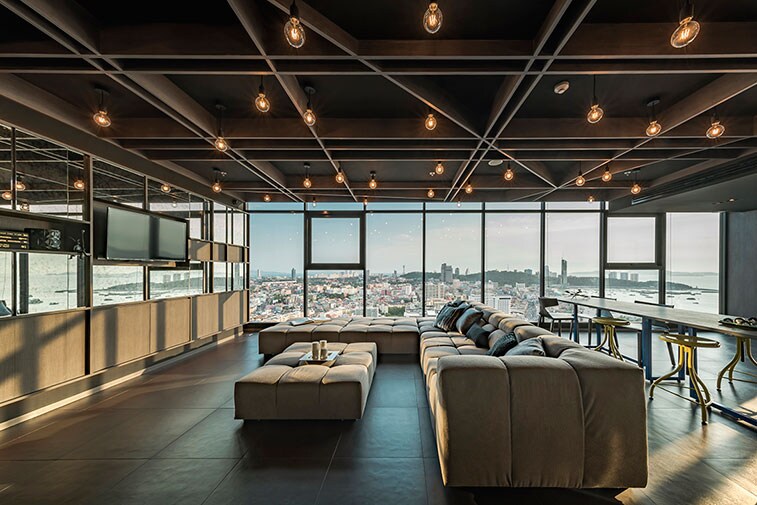 A spacious living area features panoramic floor-to-ceiling windows offering a view of the city and sea. Stylish light fixtures illuminate the room, where a large sectional sofa is complemented by a modern table and seating arrangement, creating an inviting space for relaxation.