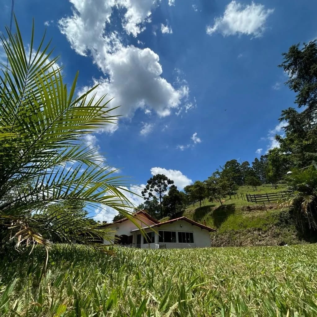 The image showcases a charming house surrounded by lush greenery under a bright blue sky with scattered clouds. Tall grass occupies the foreground, while hillside trees frame the background, highlighting the natural landscape.