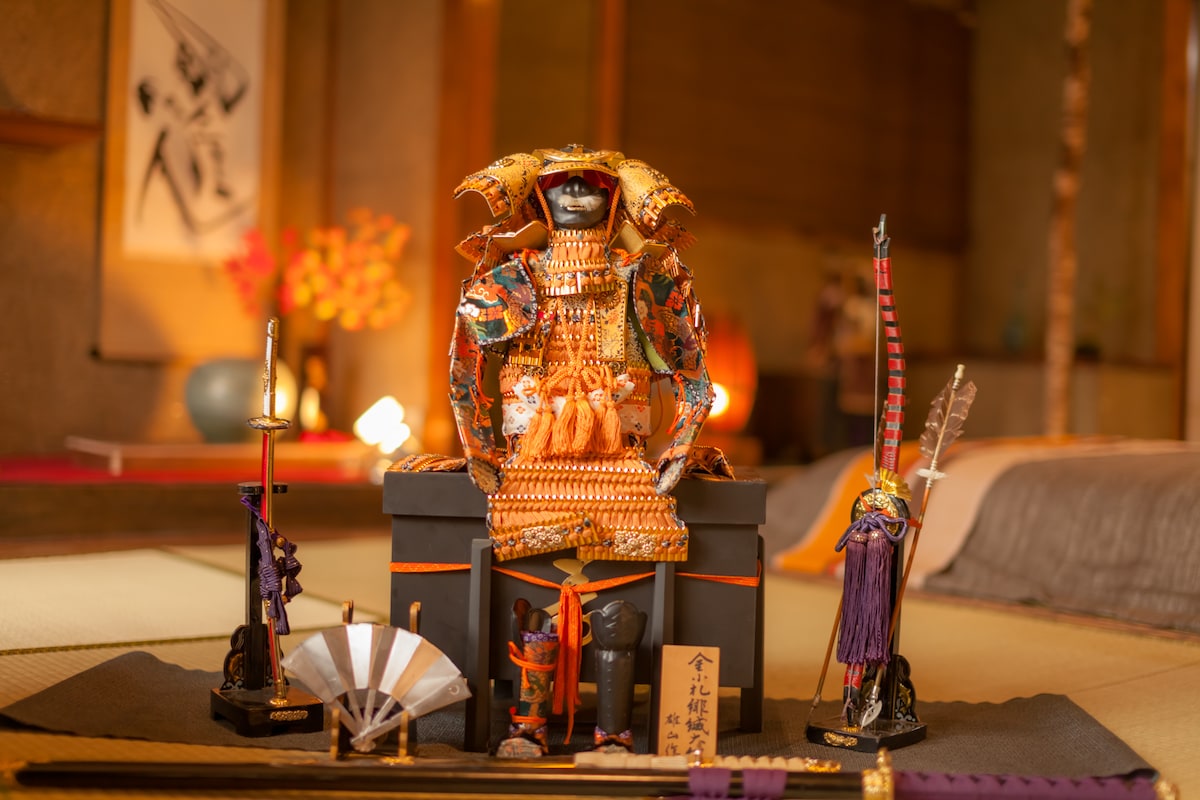 A traditional Japanese display featuring a samurai figurine adorned in intricate armor. The figure is positioned on a black pedestal alongside decorative swords, a fan, and a small purple cloth. The background includes elements of tatami flooring and softly lit furnishings, reflecting Japanese aesthetics.