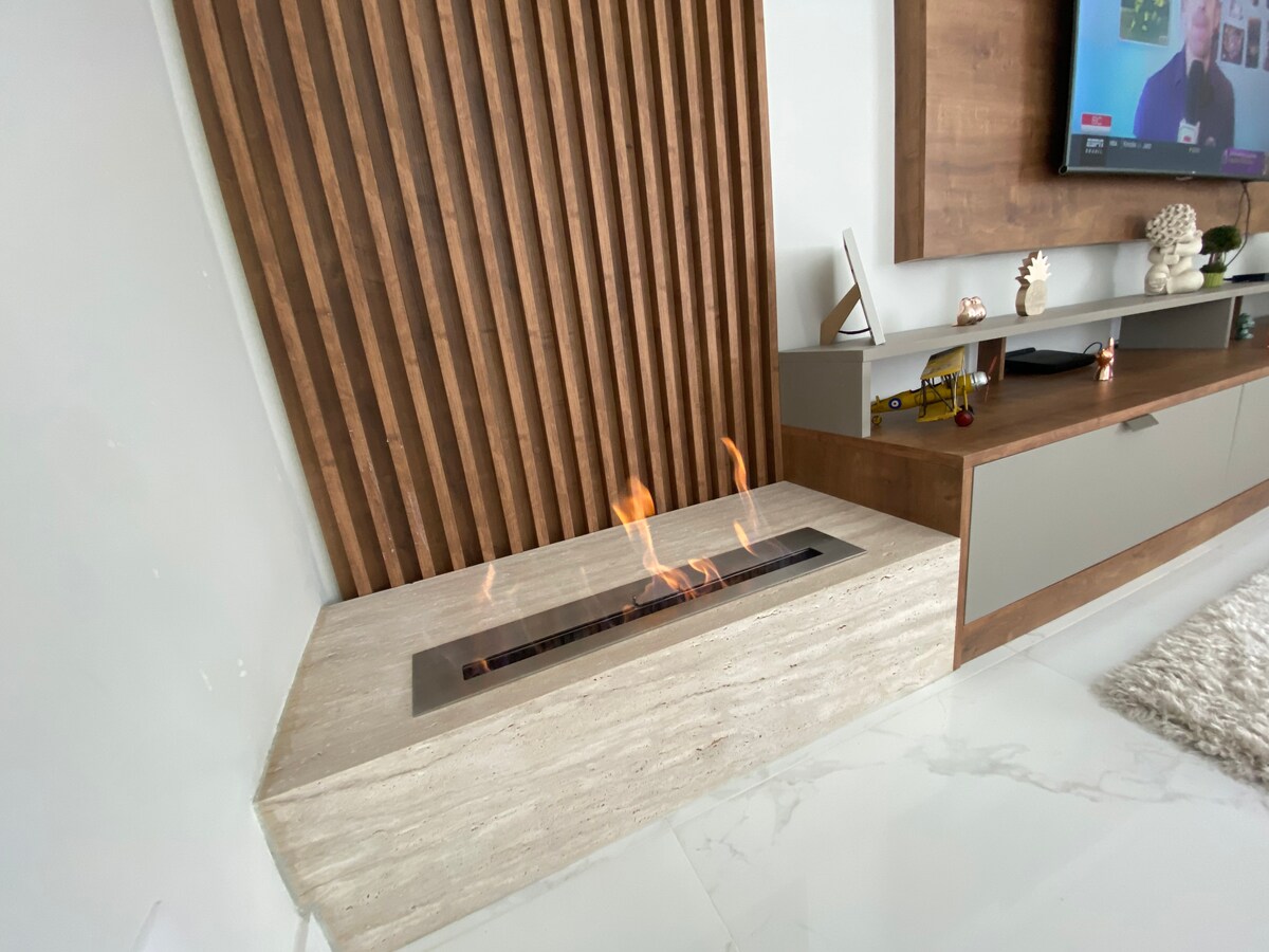 A modern fireplace is featured in a minimalist setting, with a sleek stone base and a long, rectangular flame. The background includes a wooden panel, contributing to a contemporary aesthetic, while a flat-screen television is mounted above a stylish media unit.