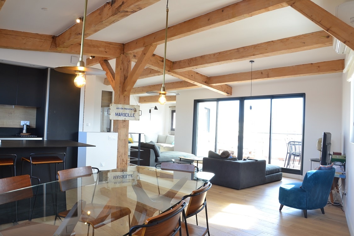 An open-plan living area features exposed wooden beams and large glass doors leading to a terrace. A dining table with several chairs is positioned near the kitchen, while a spacious seating area with a sofa invites relaxation.